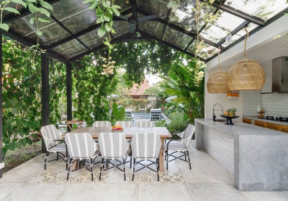 Open kitchen with empty dining room table and chairs outside, against green fresh plants on background